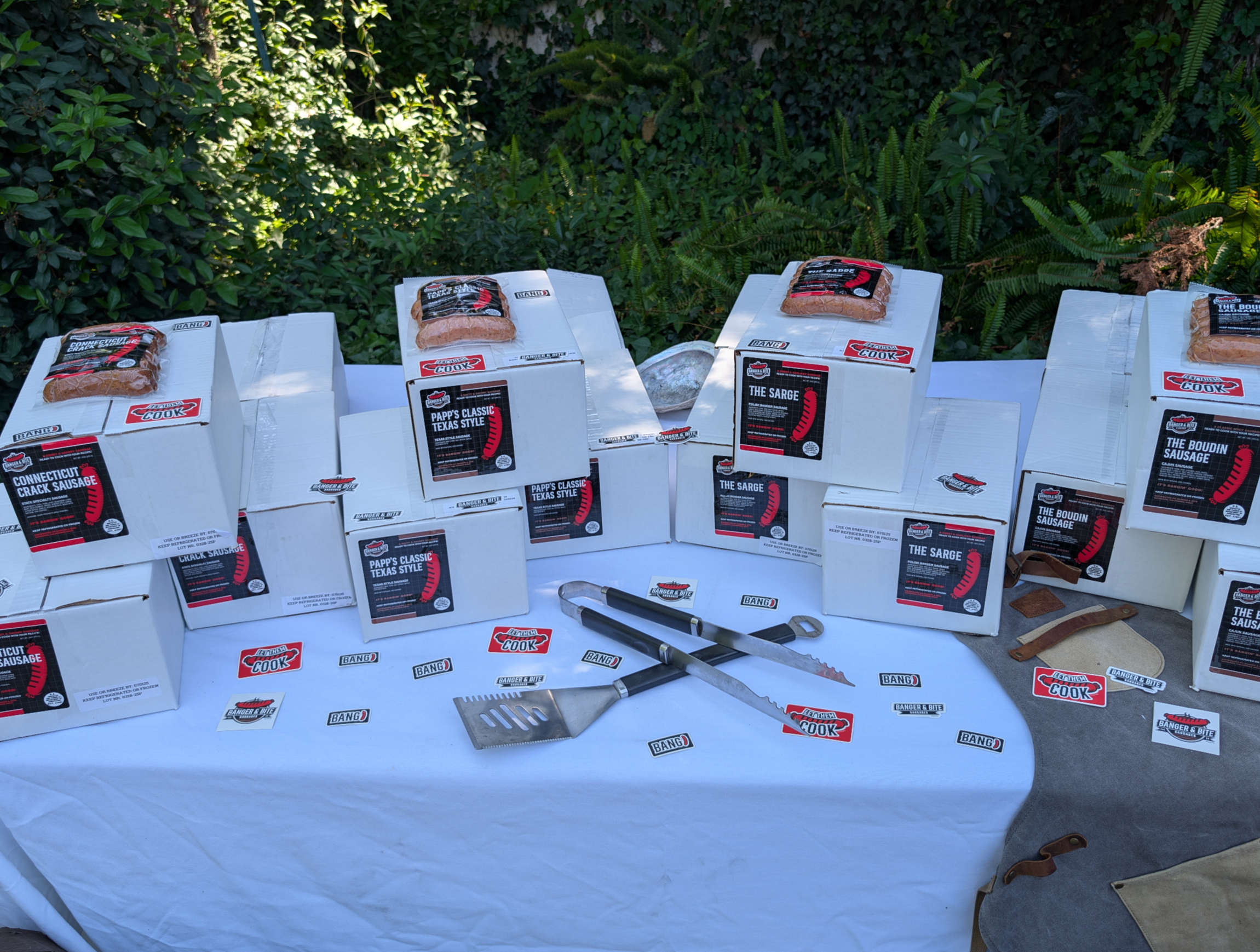 Collection of packaged sausages in boxes and grilling tools on top of a white tablecloth outdoors with greenery in the background