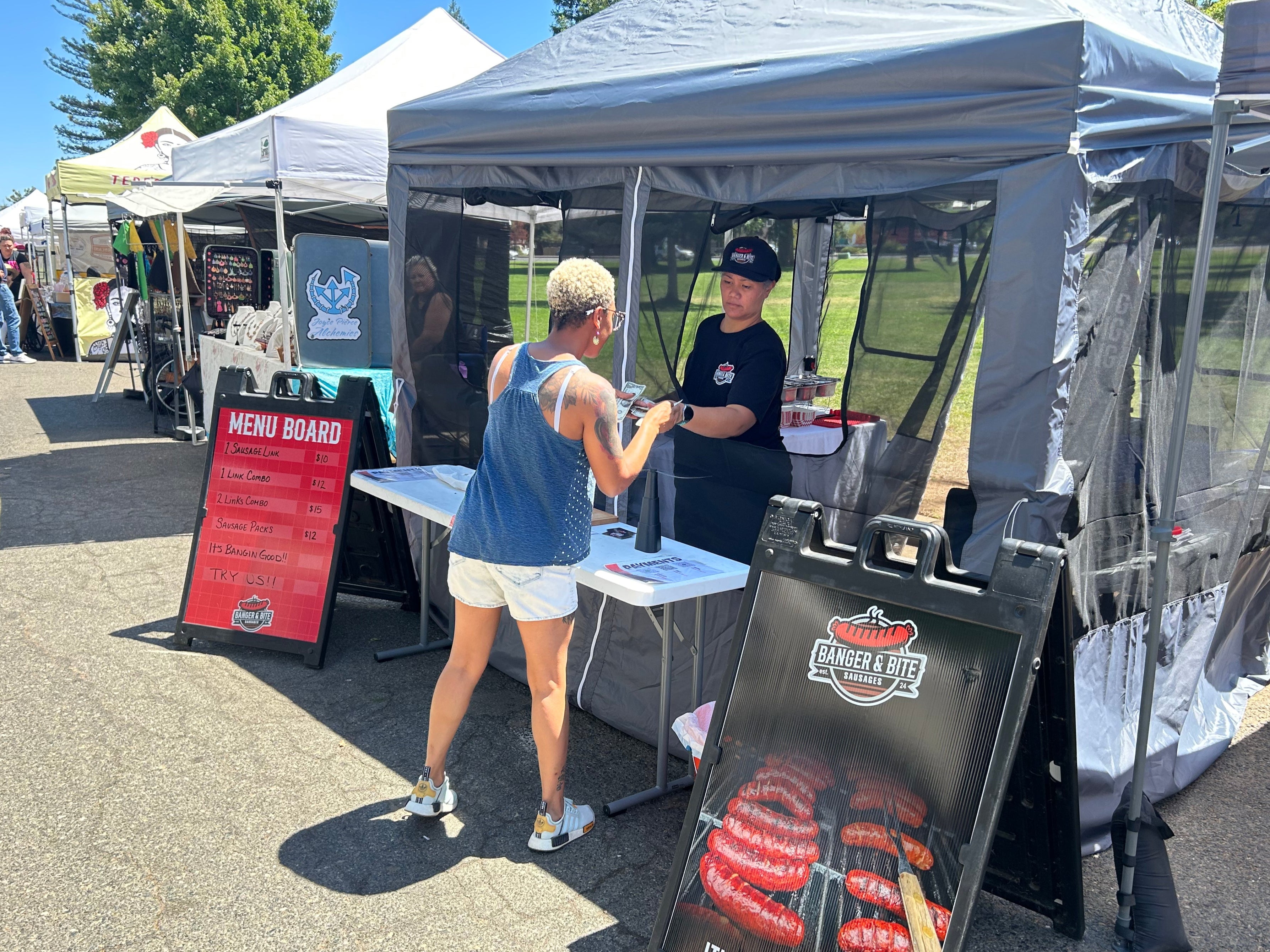 Customer ordering at our Banger & Bite food stand at a local farmer's market with a menu board and promotional sign propped up in front of it