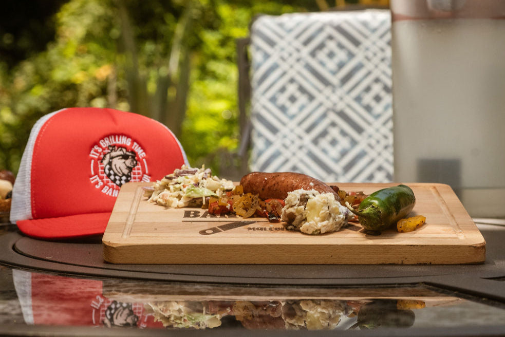 Cut up sausage with cold slaw, peppers, jalapeños, and onions on top of an engraved, wooden cutting board next to a red cap in the background