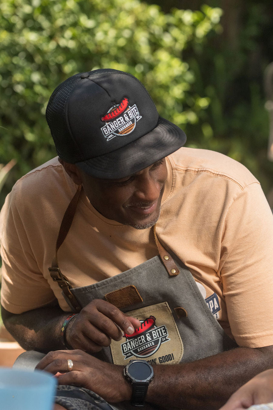 Our founder, Malcolm, wearing a Banger & Bite black cap and branded apron, sitting outdoors